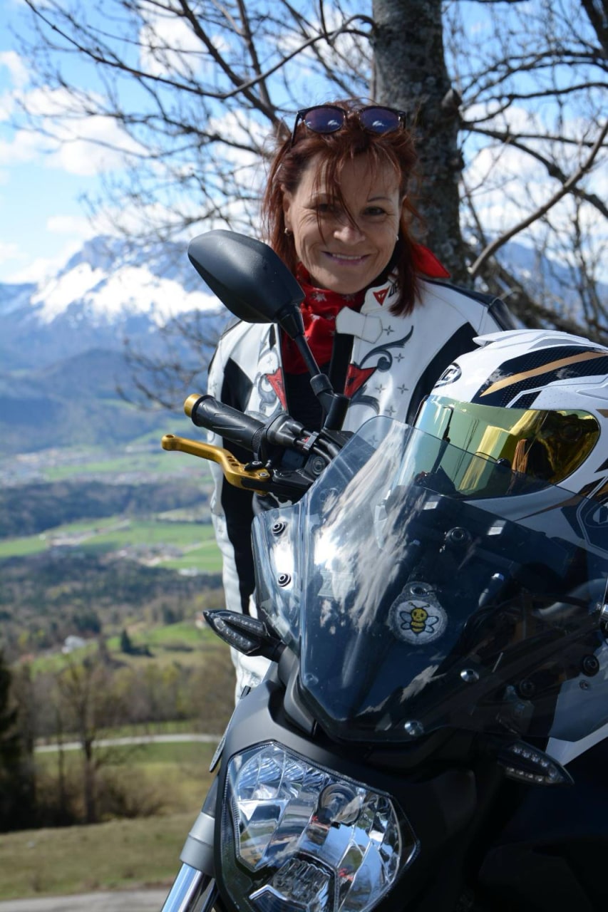 Irene Schmid in einer Motorradjacke steht während einer geführten Motorradreise neben ihrem Motorrad, mit einer malerischen Kulisse aus Bergen und Tälern im Hintergrund. Sie lächelt, die Sonnenbrille sitzt auf ihrem Kopf, während ein kahler Baum die malerische Aussicht noch verstärkt.