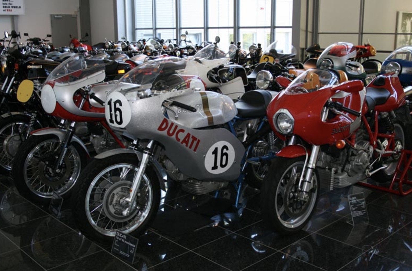 Innenausstellung von Oldtimer-Motorrädern: silberne Ducati Nr. 16, rote MV Agusta, alle auf einem glänzenden schwarzen Boden.